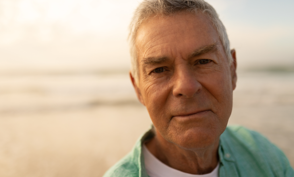 An elderly man on a beach looking down the camera lens
