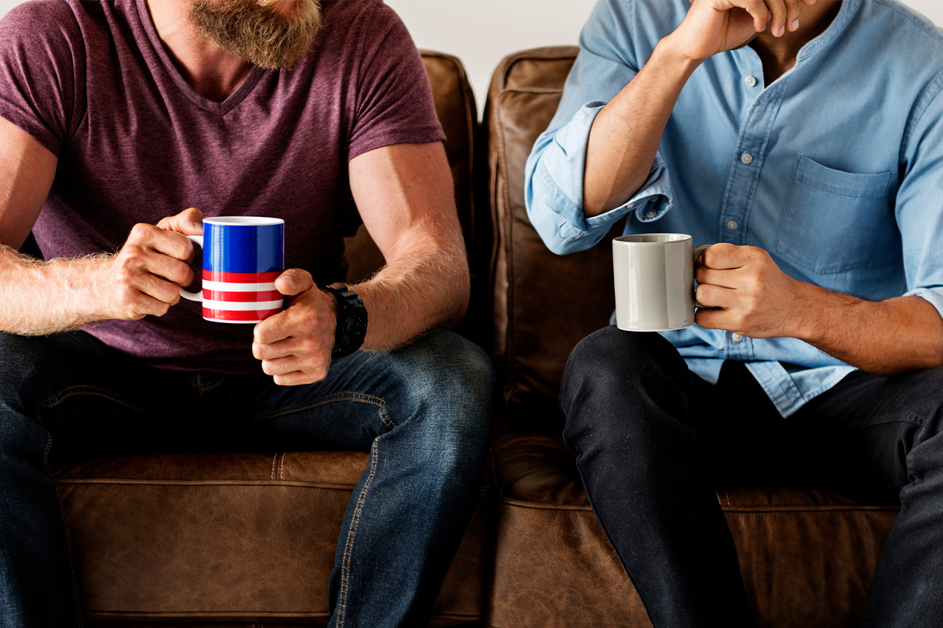 Two men sat down on a sofa with hot drinks 